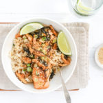 Grilled Salmon served over veggies and brown rice with an Asian soy ginger sauce and lime wedges served in a white bowl with a small bowl of sesame seeds and a glass of sparkling water.