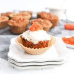 One carrot cake muffin with cream cheese icing atop 3 plates with more muffins in the background on a grey napkin.