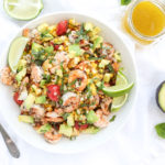 Grilled shrimp salad in a white bowl on a white linen napkin with lime wedges and dressing in a mason jar.