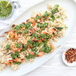 An overhead shot of grilled shrimp skewers over rice with chimichurri on a white oval plate with a jar of sauce and bowl of red pepper flakes on the side.