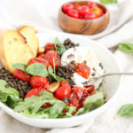 a bowl of roasted tomatoes with black lentils and toasted baguette slices with a fork cutting through the burrata